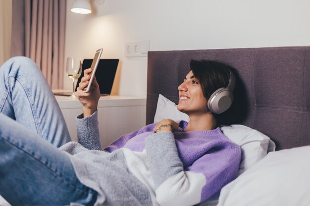 Mujer feliz en casa en una cómoda cama vistiendo suéter de ropa de abrigo, escuchar música