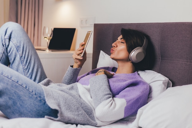 Mujer feliz en casa en una cómoda cama vistiendo suéter de ropa de abrigo, escuchar música