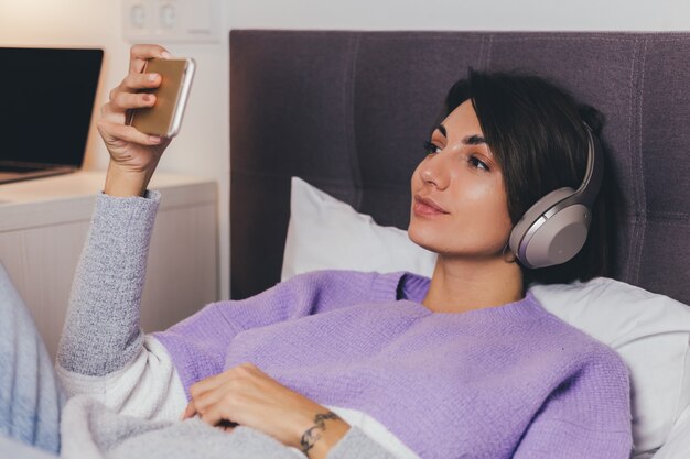 Mujer feliz en casa en una cómoda cama vistiendo suéter de ropa de abrigo, escuchar música