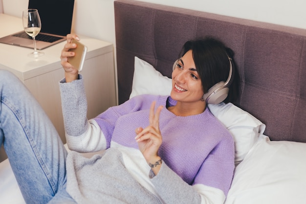 Mujer feliz en casa en una cómoda cama vistiendo suéter de ropa de abrigo, escuchar música