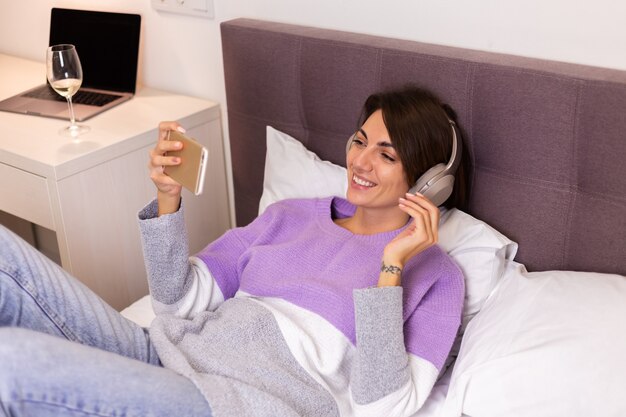 Mujer feliz en casa en una cómoda cama vistiendo suéter de ropa de abrigo, escuchar música