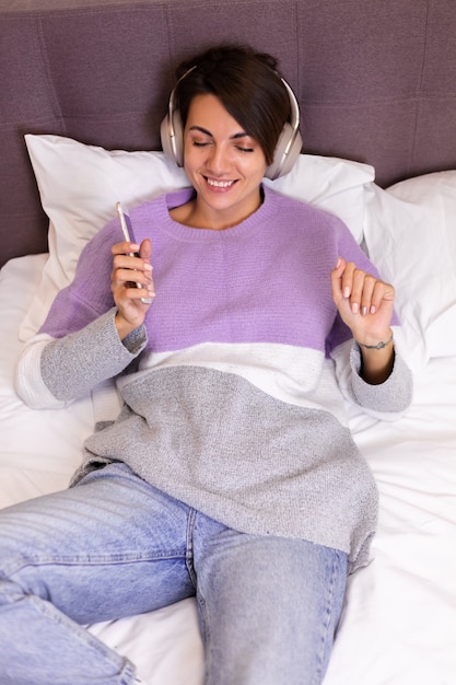 Mujer feliz en casa en una cómoda cama vistiendo suéter de ropa de abrigo, escuchar música