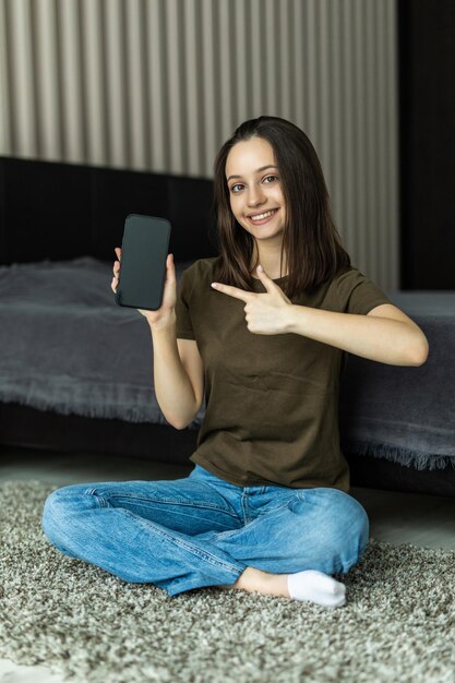 Mujer feliz en casa sentada sobre una alfombra, sosteniendo el teléfono inteligente apuntado en la pantalla