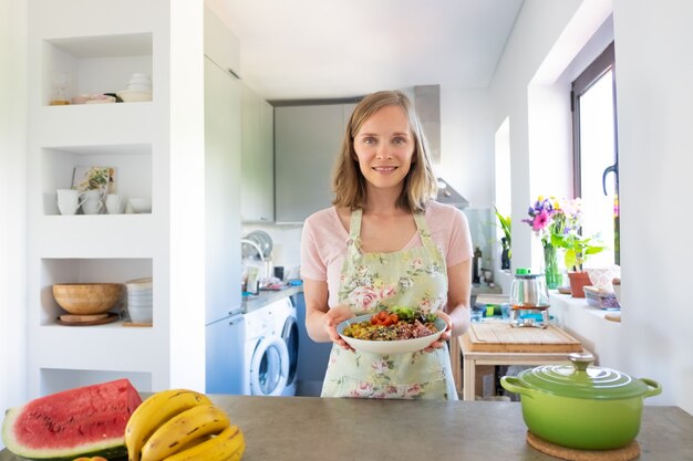 Mujer feliz cocinando en casa, manteniendo una dieta saludable, sosteniendo un plato de verduras caseras, sonriendo a la cámara. Vista frontal. Concepto de alimentación saludable