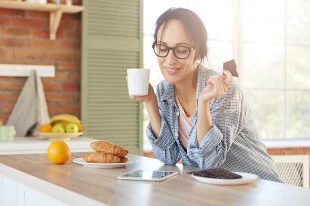 Mujer feliz come chocolate dulce y bebe té, mira una película divertida en tableta utiliza conexión a Internet de alta velocidad en casa