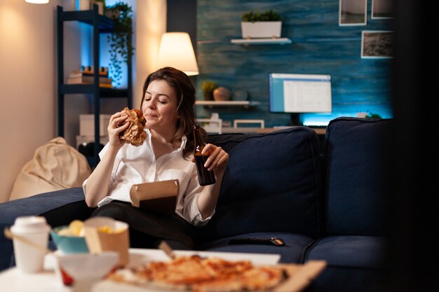 Mujer feliz comiendo deliciosa hamburguesa de entrega deliciosa relajándose en el sofá viendo una película de comedia