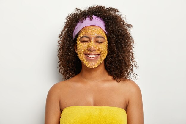 Mujer feliz y complacida con corte de pelo afro hace un exfoliante facial natural de sal marina amarilla, suaviza la piel, elimina la irritación y las manchas oscuras, mejora el equilibrio mineral, tiene una rutina de belleza, envuelto en una toalla