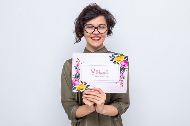 Mujer feliz y complacida con el pelo corto que sostiene la tarjeta de felicitación mirando a la cámara sonriendo alegremente celebrando el día internacional de la mujer el 8 de marzo de pie sobre fondo blanco.