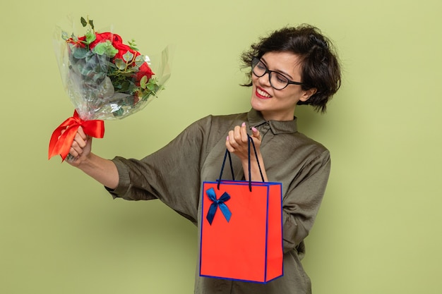 Mujer feliz y complacida con el pelo corto con ramo de flores y bolsa de papel con regalos sonriendo alegremente