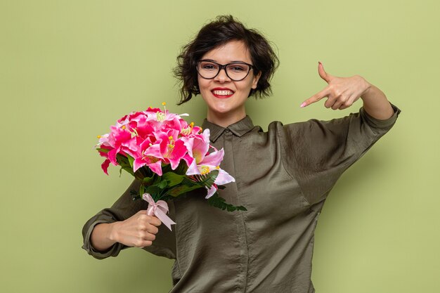 Mujer feliz y complacida con el pelo corto sosteniendo un ramo de flores apuntando con el dedo índice sonriendo alegremente celebrando el día internacional de la mujer el 8 de marzo de pie sobre fondo verde