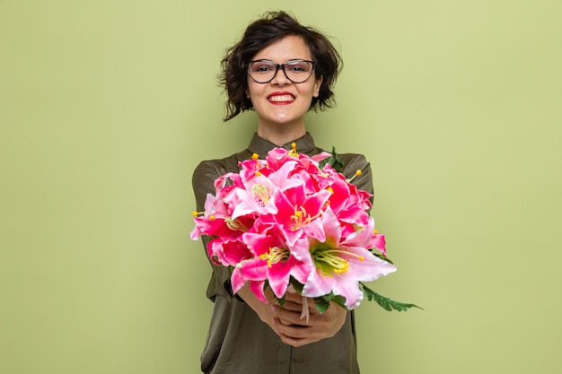 Mujer feliz y complacida con el pelo corto sosteniendo un ramo de flores mirando a la cámara sonriendo alegremente celebrando el día internacional de la mujer el 8 de marzo de pie sobre fondo verde