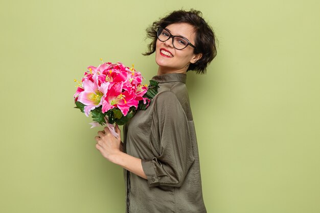 Mujer feliz y complacida con el pelo corto sosteniendo un ramo de flores mirando a la cámara sonriendo alegremente celebrando el día internacional de la mujer el 8 de marzo de pie sobre fondo verde