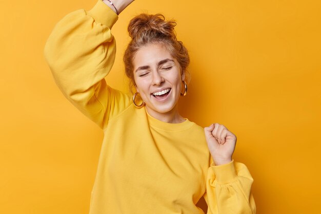 Una mujer feliz y despreocupada baila el triunfo celebra el premio ganador se ríe alegremente mantiene los ojos cerrados vestida con una sudadera casual se mueve contra el fondo amarillo se divierte Hurra dulce éxito