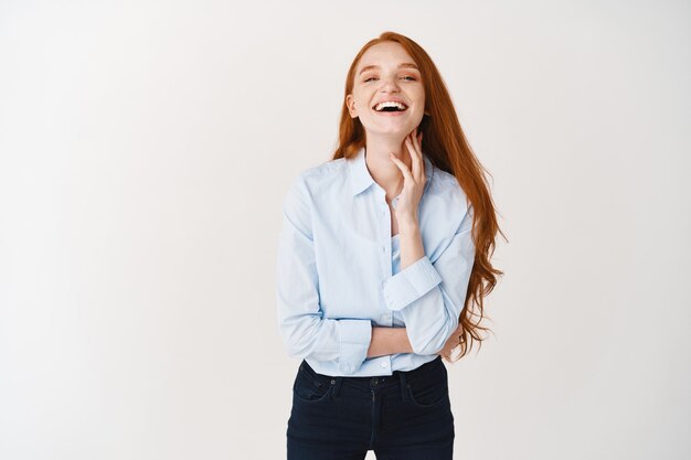 Mujer feliz y despreocupada con largo cabello rojo natural riendo, expresa alegría y felicidad, de pie contra la pared blanca