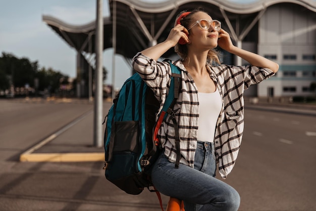 Mujer feliz disfruta del clima soleado y usa auriculares rojos Chica escucha música afuera Turista en jeans y camisa a cuadros sostiene mochila