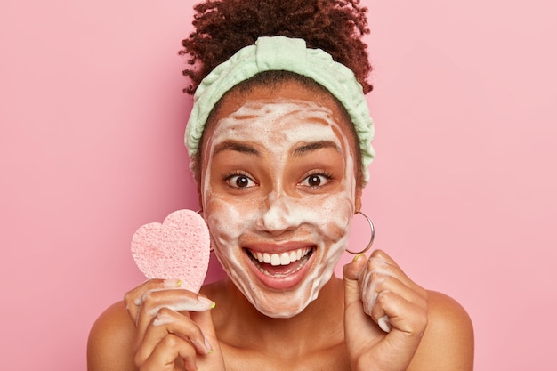 La mujer feliz disfruta de un tiempo de relajación, se lava la cara con pompas de jabón, se siente fresca y encantada, sostiene una esponja cosmética para limpiar la tez