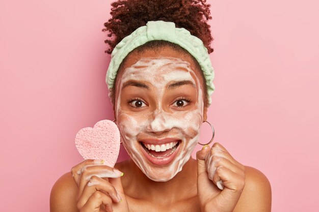 La mujer feliz disfruta de un tiempo de relajación, se lava la cara con pompas de jabón, se siente fresca y encantada, sostiene una esponja cosmética para limpiar la tez