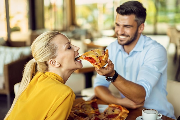 Mujer feliz divirtiéndose mientras su novio la alimenta con pizza durante el almuerzo