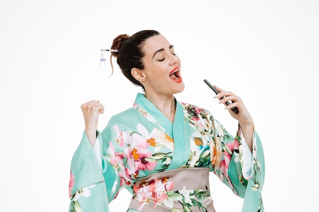 Mujer feliz y emocionada en kimono japonés tradicional con smartphone usándolo como micrófono cantando una canción divirtiéndose en blanco