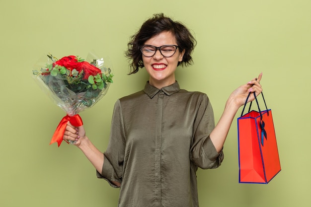 Mujer feliz y emocionada con el pelo corto con ramo de flores y bolsa de papel con regalos sonriendo alegremente