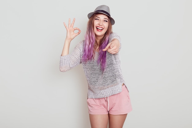 Mujer feliz emocionada con el pelo morado apuntando a la cámara con el dedo y mostrando el signo bien. gesto de aprobación, expresando emociones positivas, aislado sobre una pared blanca.