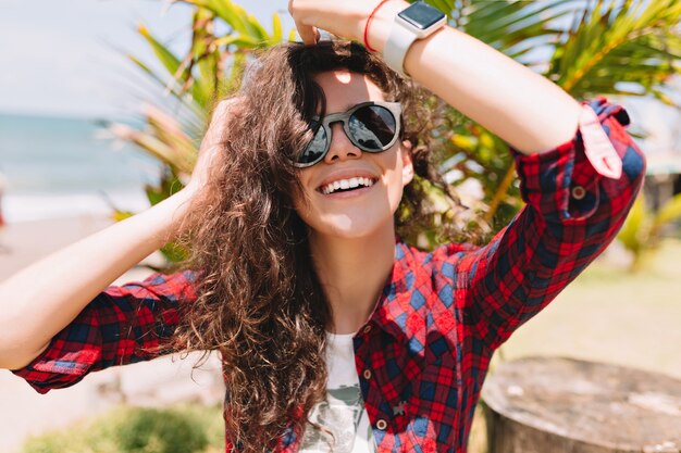 La mujer feliz emocionada con el pelo ondulado lleva gafas de sol se ve feliz y sonríe. Vacaciones de verano