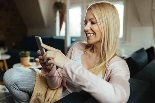 Mujer feliz enviando mensajes de texto en un teléfono inteligente mientras se relaja en la sala de estar