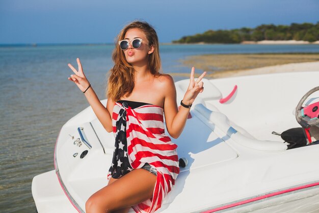 Mujer feliz envuelta en bandera americana en vacaciones tropicales de verano viajando en barco en el mar, fiesta en la playa, gente divirtiéndose juntos, emociones positivas