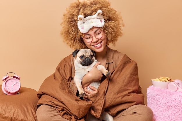 Una mujer feliz envuelta en una manta sostiene un perro pug que expresa su amor por su mascota favorita tiene poses de expresión alegres en una cómoda cama despertador tazón de copos de maíz alrededor de un fondo beige Es hora de descansar