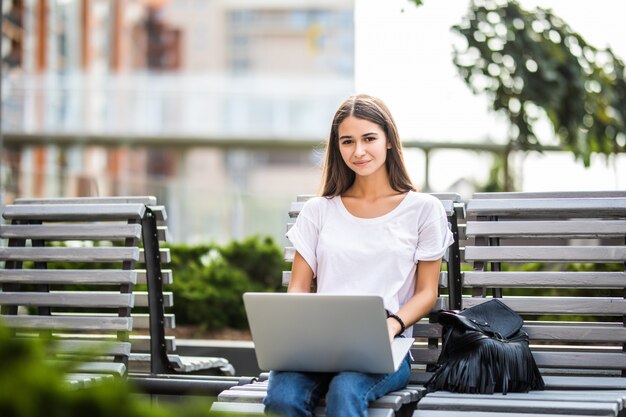 Mujer feliz escribiendo en una computadora portátil y mirando a la cámara sentada en un banco al aire libre