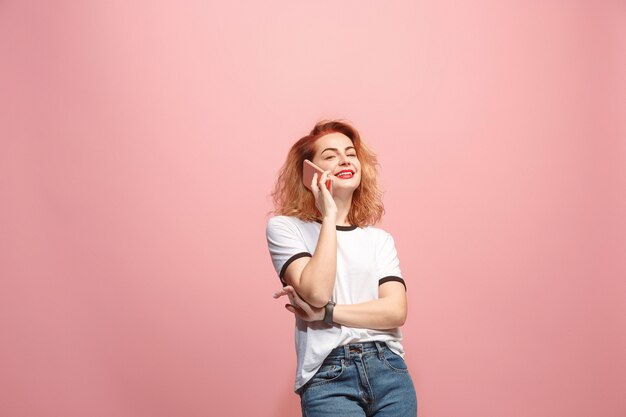 La mujer feliz está sonriendo y hablando en el teléfono inteligente contra la pared de color rosa.