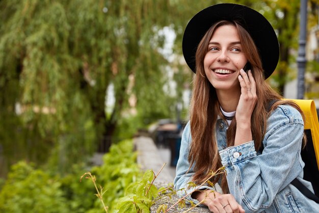 Mujer feliz con expresión alegre, vestida con elegante sombrero negro y chaqueta vaquera, llama por teléfono móvil a un amigo