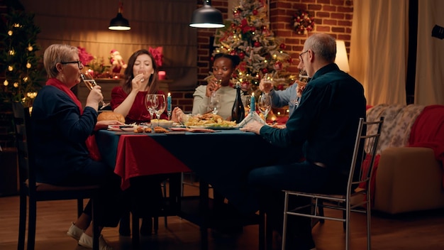 Mujer feliz festiva brindando con miembros de la familia mientras disfruta de la cena casera de Navidad en casa. Gente multiétnica alegre celebrando las tradicionales vacaciones de invierno mientras bebe vino espumoso.