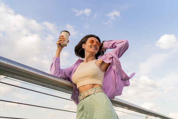 Mujer feliz con figura perfecta en elegante camisa de gran tamaño púrpura disfrutando de una taza de café mientras camina en el puente moderno