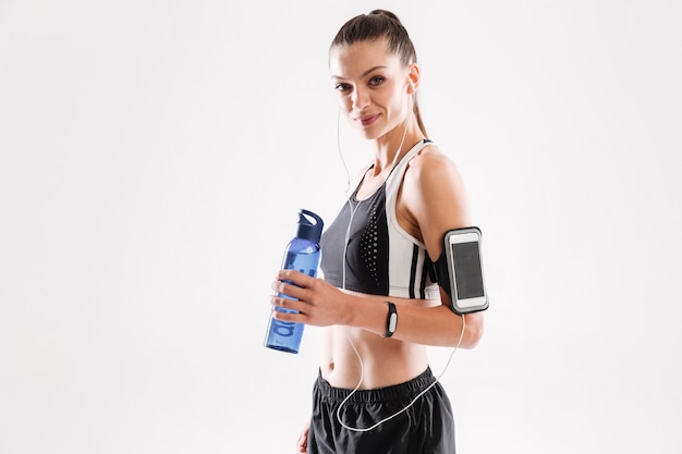 Mujer feliz fitness en auriculares de pie y sosteniendo la botella de agua