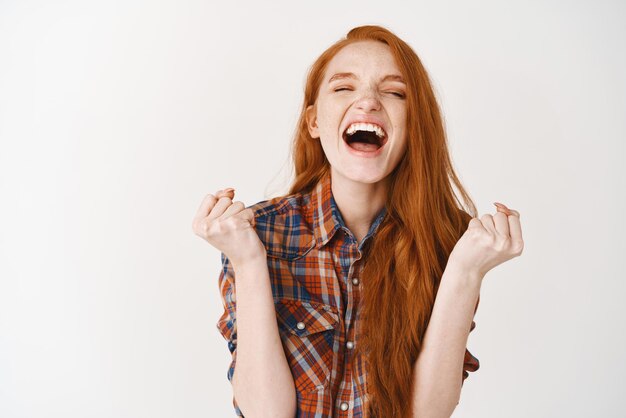 Mujer feliz ganando grito de alegría y triunfo Chica pelirroja alcanza la meta y celebra gritando de alegría de pie sobre fondo blanco