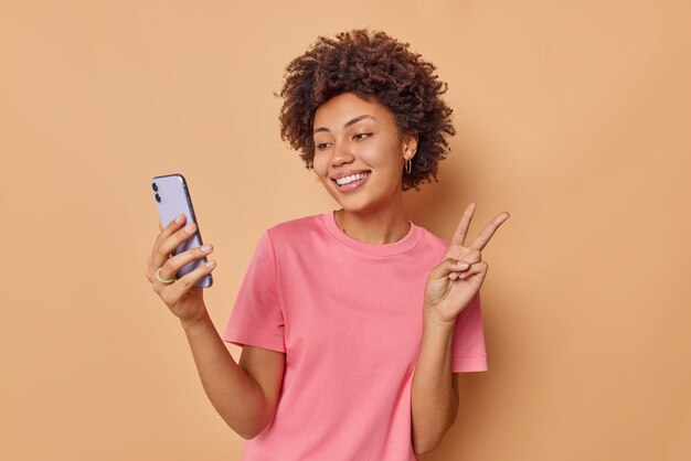 Mujer feliz con una gran sonrisa en la cara toma selfie en el teléfono móvil hace que el gesto de paz capture el momento disfruta de una conversación amistosa con un amigo a distancia vestido informalmente aislado sobre una pared beige