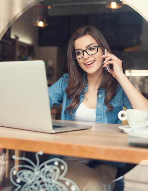 Foto gratuita mujer feliz hablando por teléfono móvil y usando la computadora portátil en el café