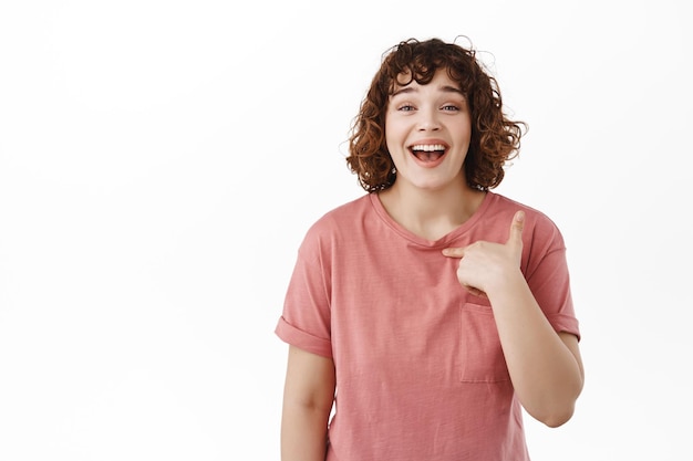 Mujer feliz y hermosa apuntándose a sí misma, riendo y sonriendo con dientes blancos y perfectos, siendo elegida, promovida o conseguida trabajo, autopromocionada o fanfarroneando, de pie sobre fondo blanco