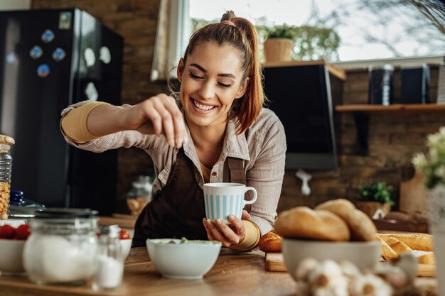 Mujer feliz joven que prepara una comida y que sazona la comida en la cocina