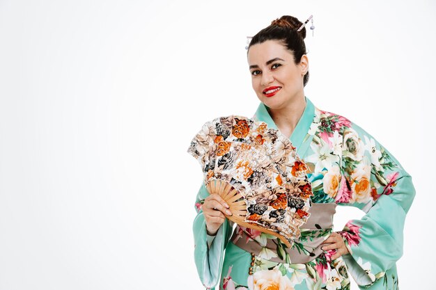 Mujer feliz en kimono japonés tradicional sosteniendo ventilador de mano sonriendo alegremente en blanco