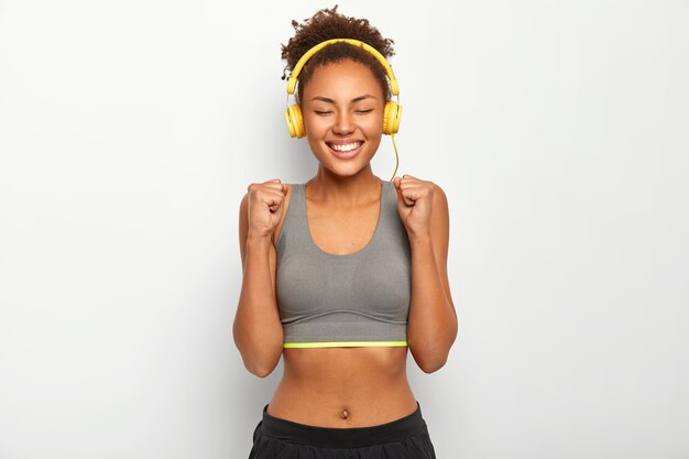 Mujer feliz levanta los puños cerrados, celebra el juego ganado con éxito, vestida con sujetador deportivo, escucha música a través de auriculares