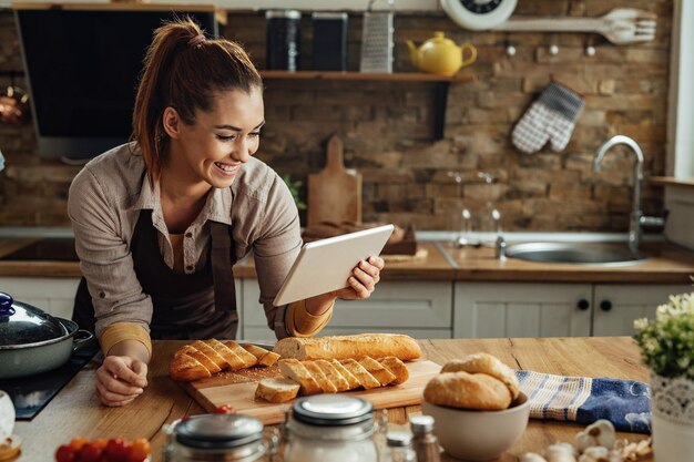 Mujer feliz leyendo la receta en el panel táctil mientras prepara la comida en la cocina.