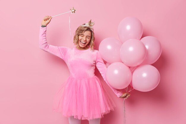 Una mujer feliz y llena de alegría con peinado viste un vestido festivo celebra una ocasión especial sostiene una varita mágica y un montón de globos de helio aislados sobre fondo rosa Concepto de personas y vacaciones