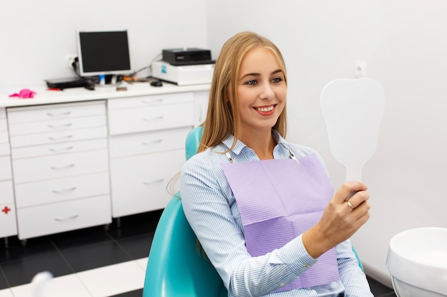 La mujer feliz mira sus dientes en el espejo que se sienta en la oficina del dentista