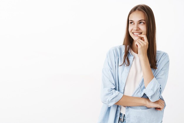 Mujer feliz mirando en la esquina superior izquierda con una sonrisa romántica satisfecha y curiosa sosteniendo el dedo en el labio sonriendo y cruzando el cuerpo con el brazo