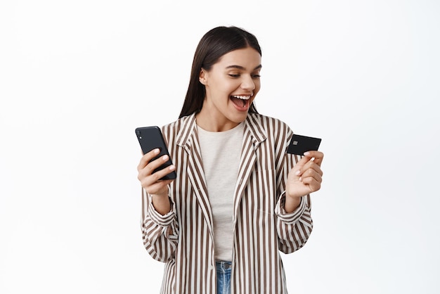 Mujer feliz mirando su tarjeta de crédito mientras usa la aplicación de compras de teléfonos inteligentes de pie con un traje elegante contra el fondo blanco