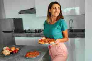 Foto gratuita mujer feliz morena que sostiene la placa con la pizza en cocina en casa moderna