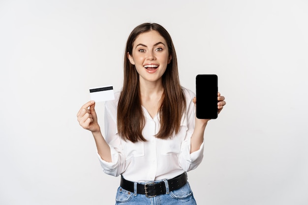 Mujer feliz mostrando tarjeta de crédito y pantalla de teléfono inteligente, concepto de compras en línea, compra en aplicación, de pie sobre fondo blanco