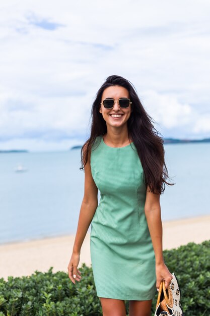 Mujer feliz muy elegante en vestido verde de verano con bolsa, con gafas de sol de vacaciones, mar azul de fondo